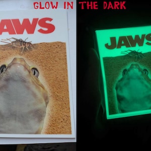 Puede incluir: Una pegatina que brilla en la oscuridad con el cartel de la película Jaws. La pegatina muestra una rana mirando hacia arriba a un grillo con la palabra "JAWS" en letras grandes sobre la rana.