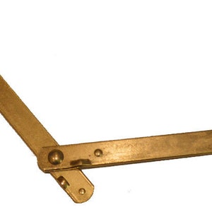 May include: A brass window stay, a type of hinge, with a flat, rectangular arm and a pivoting joint. The stay is a golden color and has a small, rounded knob at the end of the arm. The stay is designed to hold a window open.