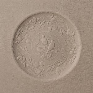 May include: A beige, circular embossed design featuring two birds perched on a branch within a circle. The circle is surrounded by floral and leaf patterns. The design is on a textured surface.