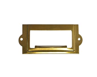 May include: A gold metal label holder with rounded corners and two small holes on the sides. The holder has a rectangular opening for inserting a label.