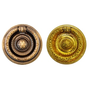 May include: Two ornate metal drawer pulls with a ring handle. One pull is brass with a gold finish and the other is brass with a dark brown finish.