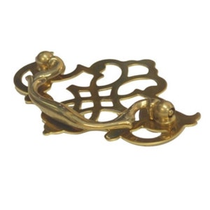 May include: A gold-toned metal cabinet pull with a decorative, ornate design. The pull has a curved handle and a decorative backplate.