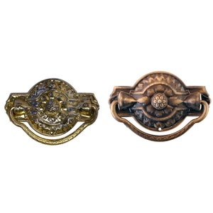 May include: Two antique drawer pulls. One is gold-toned with intricate floral and geometric patterns. The other is bronze-toned, also featuring floral designs. Both have a curved handle and are likely made of metal.