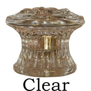 May include: Clear glass cabinet knob with a decorative floral design on top. The knob has a ribbed, hourglass-shaped body and a brass-colored metal stem. The word "Clear" is printed in black at the bottom.