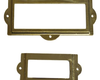 May include: Two gold-colored metal label holders. The larger holder is rectangular with rounded corners and a small lip on the top. The smaller holder is also rectangular with rounded corners.