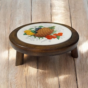 Puede incluir: Un soporte para plantas redondo de madera con una superficie de baldosas de cerámica. La baldosa presenta una colorida ilustración de piña, cerezas, limones y arándanos. El soporte mide unos 15 cm de diámetro y tiene tres patas cortas.