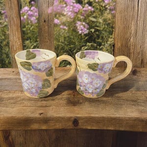 May include: Two ceramic mugs with a floral design. The mugs have a light yellow base colour with purple and white flowers and green leaves. The mugs are sitting on a wooden surface.