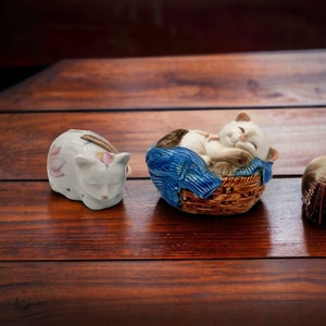 Puede incluir: Dos figuritas de cerámica de gatos. Una es blanca con flores rosas y está tumbada. La otra es blanca con detalles marrones y azules y está durmiendo en una cesta.