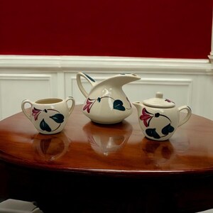 May include: A white ceramic creamer, sugar bowl, and teapot with a floral design in shades of pink, green, and blue. The set is on a wooden table.