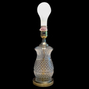 Puede incluir: Una lámpara de mesa de cristal transparente con un patrón de diamantes en la base y una base y cuello de color latón. La lámpara tiene una bombilla blanca en la parte superior. La lámpara está sobre un fondo negro.