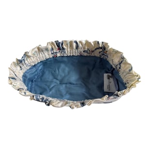 Puede incluir: Una bolsa de tela blanca con un estampado floral azul y blanco y un forro azul. La bolsa tiene un borde con volantes y una pequeña etiqueta que dice "The Laundress" en texto negro.