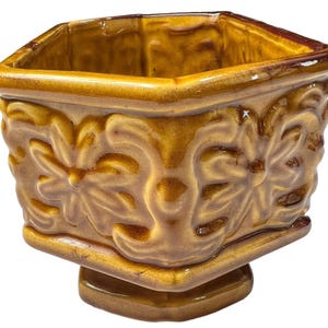 May include: A square, ceramic planter with a pedestal base. The planter is glazed in shades of brown and features a repeating floral design on the sides. The rim and base have a darker brown trim. The planter is suitable for small plants or succulents.