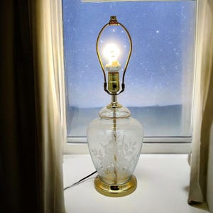 Peut inclure: Une lampe de table en verre transparent avec un motif floral gravé. La lampe a une base dorée et un abat-jour en métal doré.