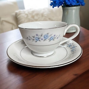 Può includere: Un set da tè bianco con un delicato motivo floreale blu. La tazza ha un bordo argentato e il piattino ha una fascia argentata attorno al bordo.