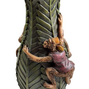 May include: A decorative vase with a leaf-like design, featuring two monkeys climbing the side. The monkeys are detailed with brown fur and wearing red and tan clothing. The vase is green and brown.