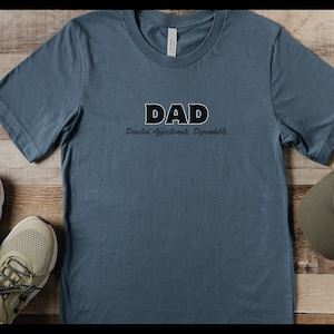 May include: A blue short-sleeve t-shirt with the word "DAD" in a bold, black and white font. Below the word "DAD" is the text "Devoted. Affectionate. Dependable." A pair of sneakers and a baseball cap are also in the image.