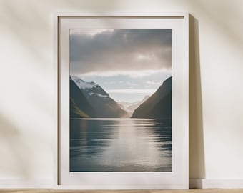 May include: A framed photograph of a mountain range with a lake in the valley. The sky is cloudy and the water is calm. The mountains are covered in green trees and snow.