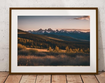 May include: A framed photograph of a mountain range with a forest in the foreground. The mountains are snow-capped and the forest is a mix of green and yellow trees. The sky is a soft pink and orange.
