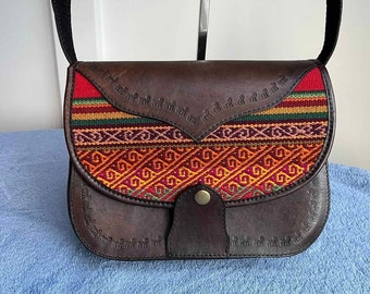 May include: A brown leather crossbody bag with a colorful woven flap. The flap features a geometric pattern in red, orange, yellow, green, and pink. The bag has a single strap and a metal button closure.