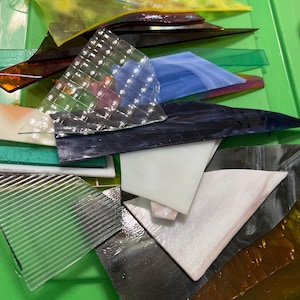 May include: A collection of colorful glass pieces in various shapes and sizes, including clear, blue, green, yellow, and brown. The glass fragments are arranged in a green plastic container, suggesting a craft or art project.