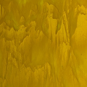 May include: A close-up of a sheet of stained glass with a flowing, textured pattern. The glass is primarily a warm, golden yellow color, with lighter, almost white, streaks and swirls throughout. The texture gives the glass a rippled appearance.