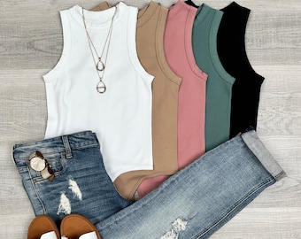 May include: Five sleeveless tops in white, beige, pink, green, and black. The tops are made of a ribbed fabric and have a mock neck. The tops are laid out on a wooden surface with a pair of distressed denim jeans and white sandals.