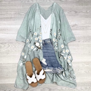 May include: A light blue floral kimono with white daisy embroidery, worn over a white v-neck top, blue denim shorts, and white sandals.