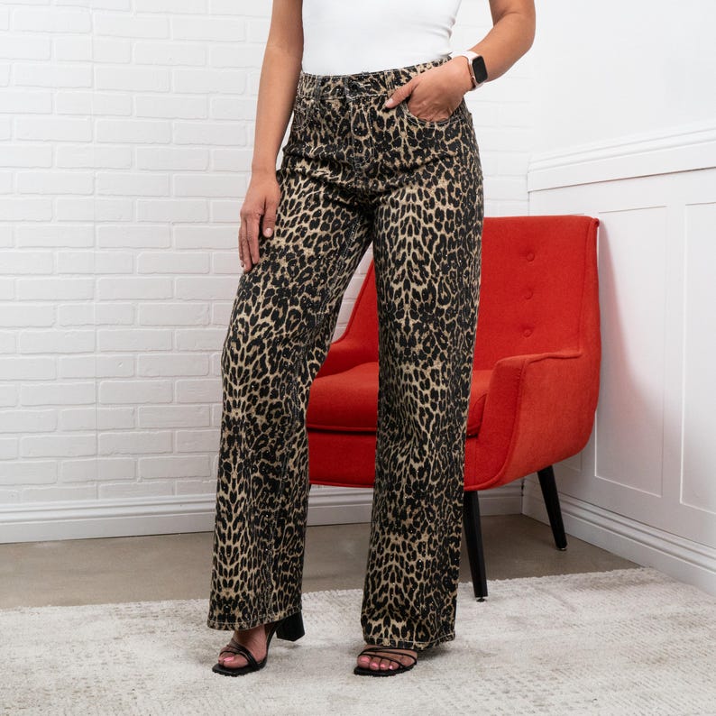 May include: Wide-leg leopard print pants in shades of black and tan. The pants have a high waist and pockets. The model is wearing black sandals. The background includes a white brick wall and a red chair.