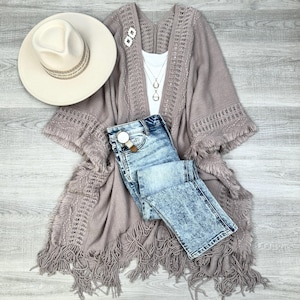May include: A mauve knitted cardigan with fringe detailing and a crocheted pattern. The cardigan is worn over a white tank top and light blue denim jeans.