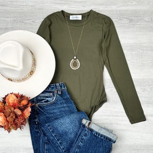 May include: A long-sleeved olive green bodysuit with a gold necklace featuring a white teardrop-shaped pendant. The bodysuit is paired with blue denim jeans and a white fedora hat with a brown trim.
