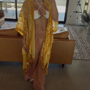 May include: A yellow kimono with a floral pattern and fringe trim, worn over a white halter-style top with a central gold-tone ring detail and light pink wide-leg trousers. The outfit is completed with white open-toe sandals and a gold-tone necklace.