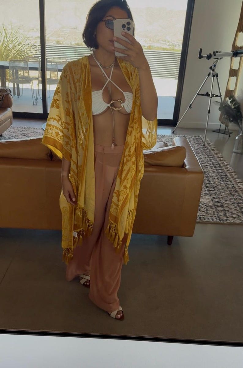 May include: A yellow kimono with a floral pattern and fringe trim, worn over a white halter-style top with a central gold-tone ring detail and light pink wide-leg trousers. The outfit is completed with white open-toe sandals and a gold-tone necklace.