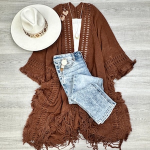 May include: A brown knitted poncho with fringe detailing, a pair of light blue denim jeans, and a white hat with brown trim.