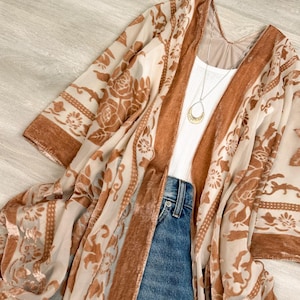 May include: A sheer kimono featuring a burnt orange velvet floral pattern, worn over a white ribbed tank top and blue denim jeans. A gold-toned teardrop pendant necklace hangs from a thin chain, resting against the white fabric of the tank top.