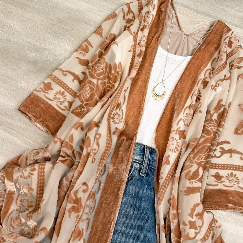 May include: A sheer kimono featuring a burnt orange velvet floral pattern, worn over a white ribbed tank top and blue denim jeans. A gold-toned teardrop pendant necklace hangs from a thin chain, resting against the white fabric of the tank top.