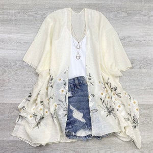 May include: A white embroidered kimono with a floral pattern of daisies. The kimono has short sleeves and a relaxed fit. It is paired with a white tank top and denim shorts.