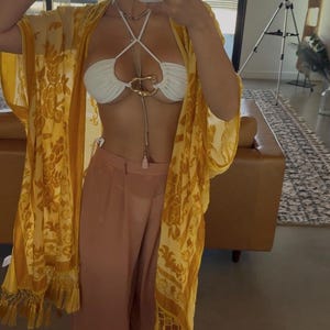 May include: A white halter bikini top featuring a central gold-toned metal ring detail, paired with loose-fitting peach-colored trousers and a sheer yellow kimono-style cover-up with a floral pattern and fringe trim along the bottom hem.