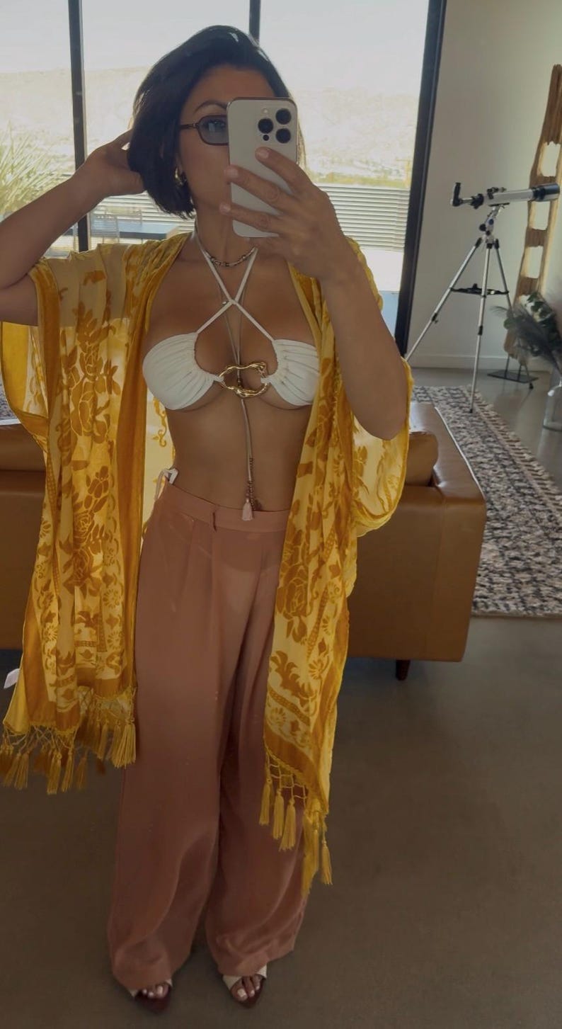 May include: A white halter bikini top featuring a central gold-toned metal ring detail, paired with loose-fitting peach-colored trousers and a sheer yellow kimono-style cover-up with a floral pattern and fringe trim along the bottom hem.