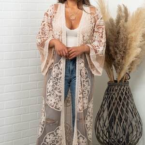 May include: A long, sheer, beige lace kimono with floral patterns and wide sleeves. The kimono is open in the front and worn over a white top and blue jeans. The background includes a white brick wall and a decorative vase.