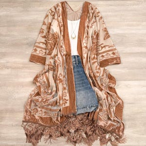 May include: A long, open-front kimono featuring a sheer, light beige fabric with a burnt orange velvet floral pattern. The garment has wide sleeves, a fringed hem, and is layered over a white tank top, blue denim shorts, and a gold pendant necklace.