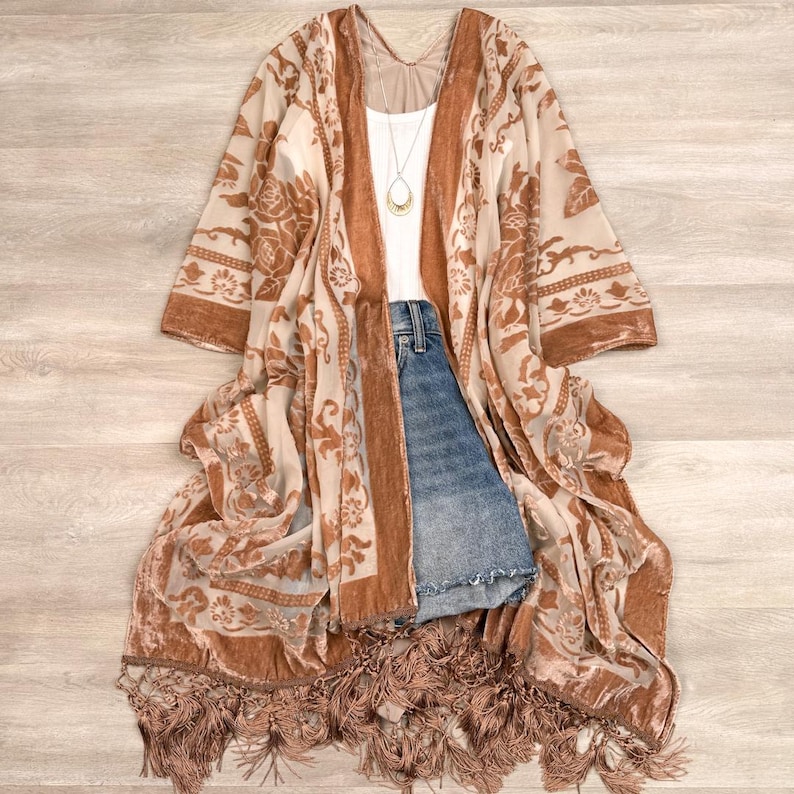 May include: A long, open-front kimono featuring a sheer, light beige fabric with a burnt orange velvet floral pattern. The garment has wide sleeves, a fringed hem, and is layered over a white tank top, blue denim shorts, and a gold pendant necklace.