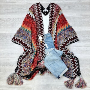 May include: A colorful knitted poncho with fringe tassels. The poncho has a geometric pattern in shades of red, black, blue, and yellow. The poncho is paired with a white tank top and blue denim shorts.