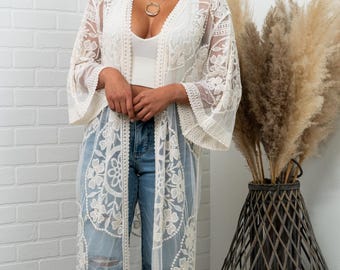 May include: A long, cream-colored lace kimono with floral patterns and wide sleeves. The kimono is worn over a white top and blue jeans. A gold necklace is visible. The background includes a white brick wall and a decorative vase with tall, dried plants.