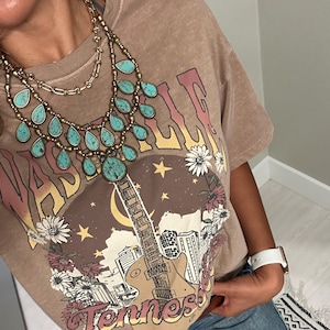 May include: A brown t-shirt with a graphic print of a guitar, city skyline, and the text "Nashville Tennessee". The shirt features a turquoise and gold layered necklace with teardrop-shaped turquoise stones.