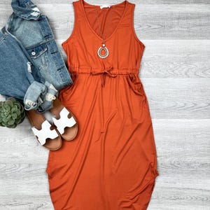 May include: A rust-colored maxi dress with a v-neck, sleeveless, and a drawstring waist. The dress has pockets on the sides and a relaxed fit.