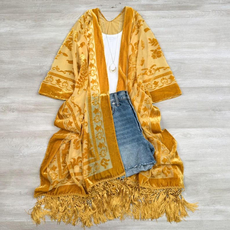 May include: A mustard yellow velvet kimono featuring a floral pattern, long sleeves, and a long fringe hem. The kimono is layered over a plain white tank top with a thin silver necklace and a pair of blue denim shorts with a button fly closure.