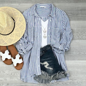 May include: A casual summer outfit featuring a straw hat, blue and white striped button-down shirt, white tank top, denim shorts, sandals, and a necklace. The shirt is rolled up at the sleeves, and the shorts have distressed detailing.