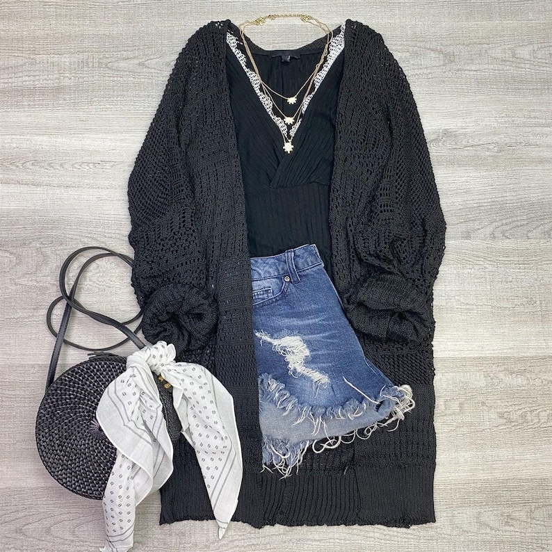 May include: A black crochet cardigan with a long, loose fit. The cardigan has a V-neck and long sleeves. It is paired with a black tank top, denim shorts, and a white bandana.