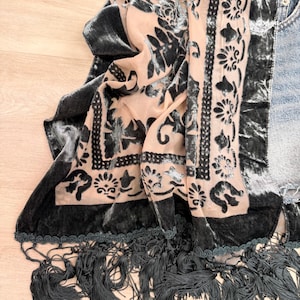 May include: A rectangular scarf featuring a sheer peach-colored base with a dark gray velvet floral and geometric pattern. The edges are finished with a dark gray velvet border and a long, thick fringe trim along the bottom hem.