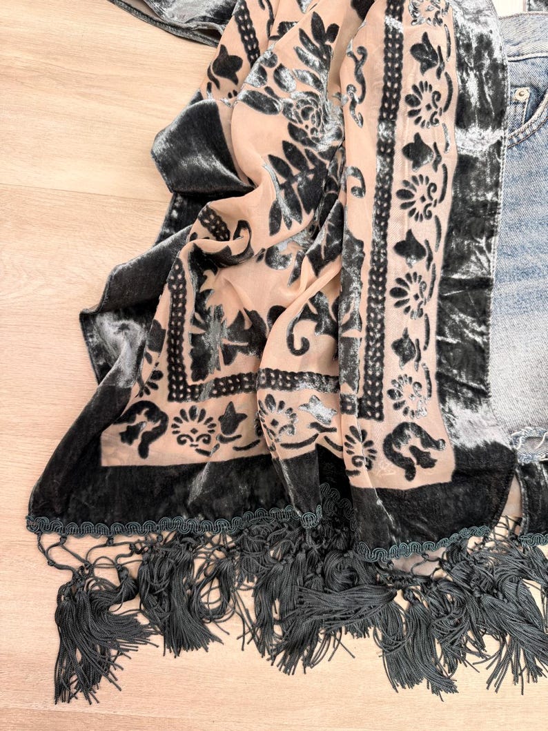 May include: A rectangular scarf featuring a sheer peach-colored base with a dark gray velvet floral and geometric pattern. The edges are finished with a dark gray velvet border and a long, thick fringe trim along the bottom hem.